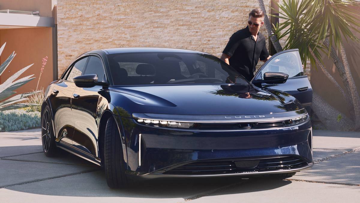 A sleek blue Lucid Air electric car is parked in a modern driveway, with a person stepping out and tropical plants in the background.