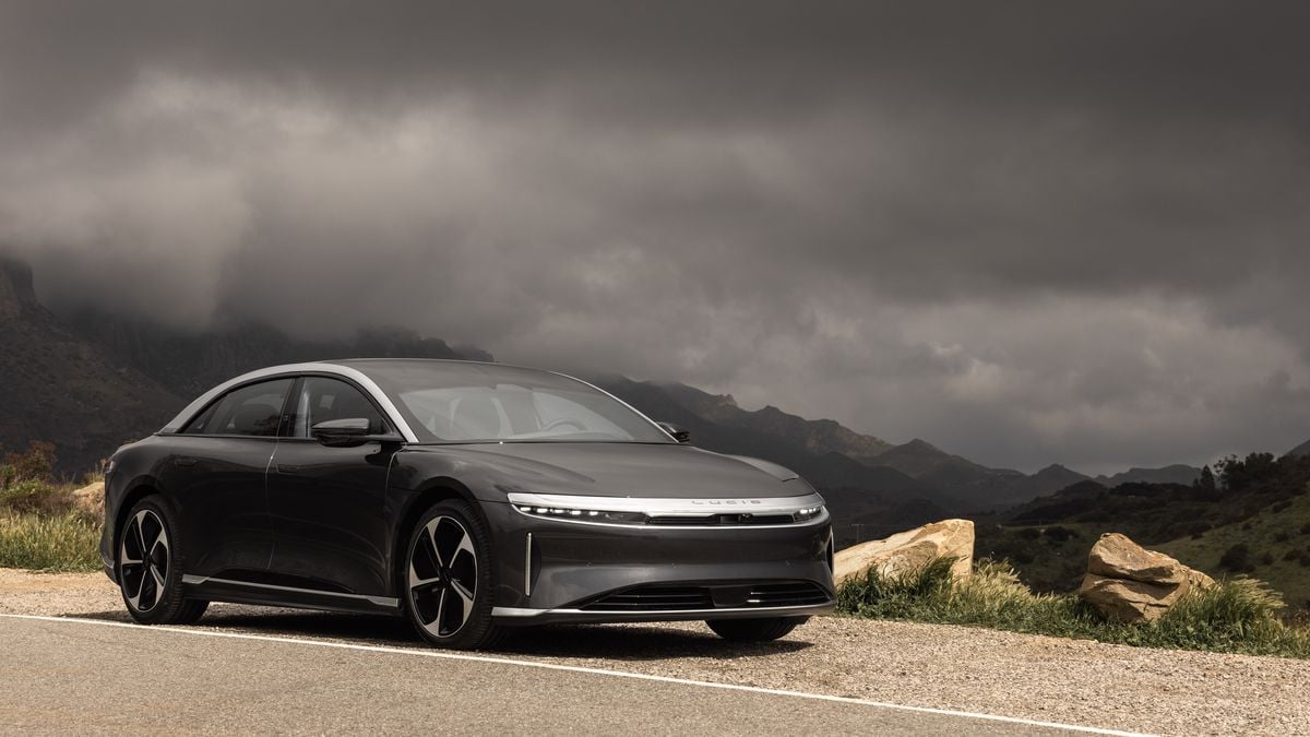 A sleek, silver electric car parked on a winding mountain road under dramatic gray clouds, surrounded by rugged landscapes.