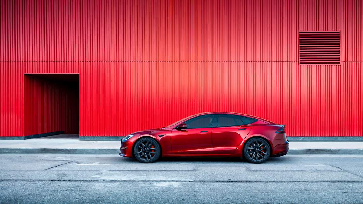 A red 2025 Tesla Model S car parked along a textured red wall, showcasing a modern urban aesthetic and vibrant color contrast.