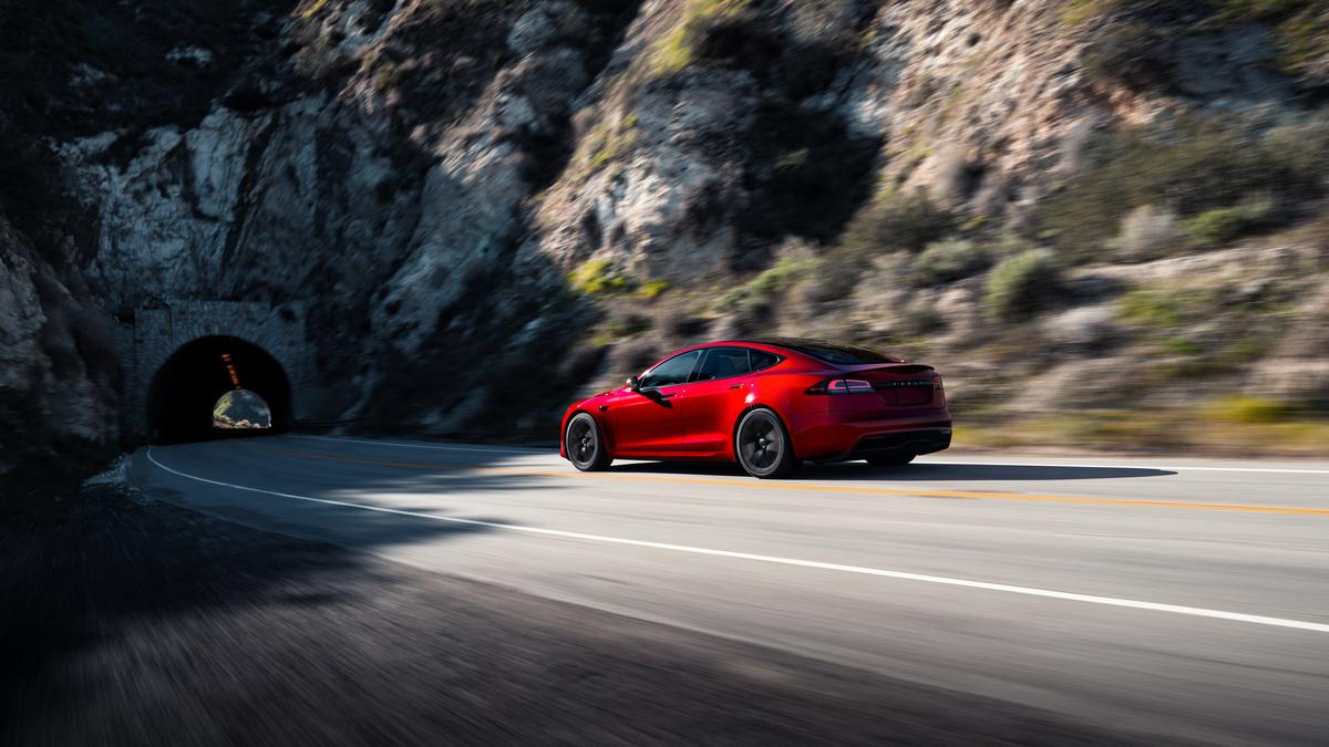 A 2025 Tesla Model S car speeds along a curvy road, nearing a dark tunnel surrounded by rocky terrain and greenery.
