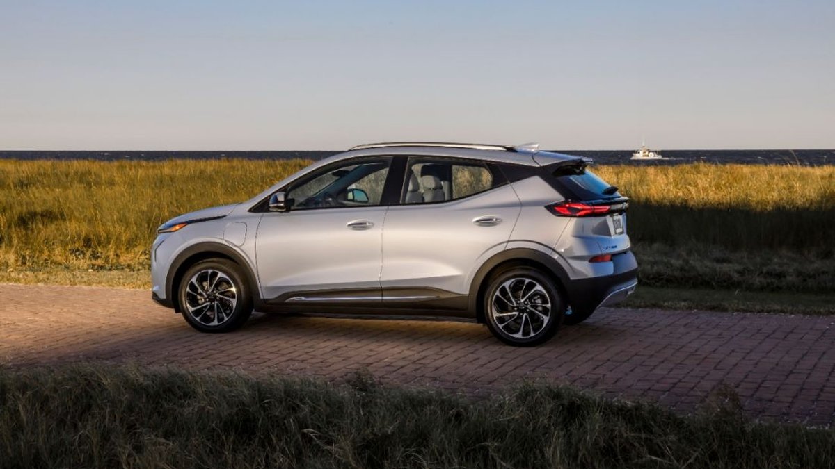 A silver electric SUV parked on a paved road, with tall grass and a calm sea in the background under a clear sky.