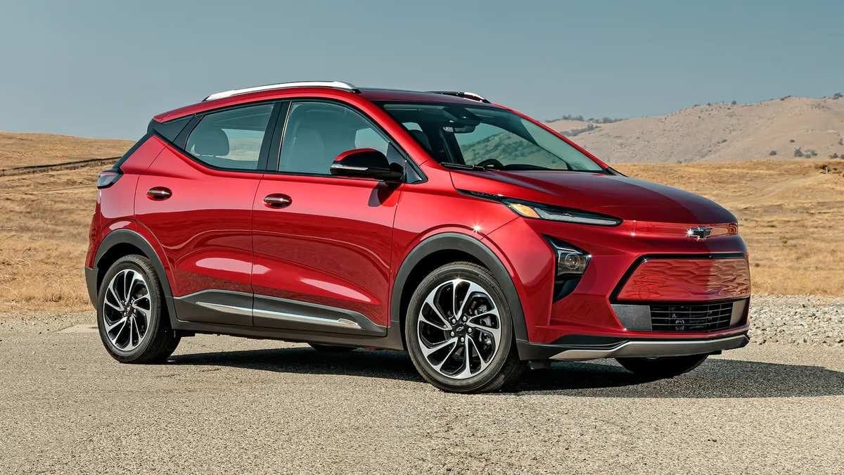 A shiny red Chevrolet Bolt EV parked on a gravel road, with a dry, hilly landscape in the background under a clear blue sky.