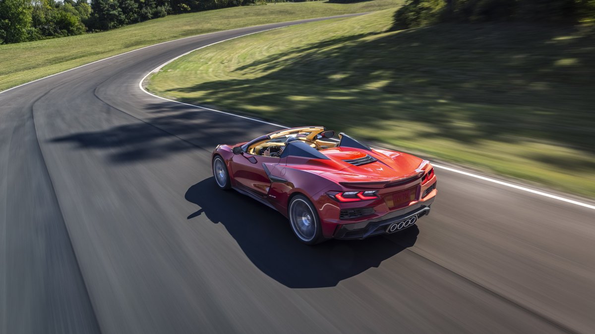 A red C8 Corvette Z06 navigates a curving racetrack surrounded by lush greenery under bright sunlight.