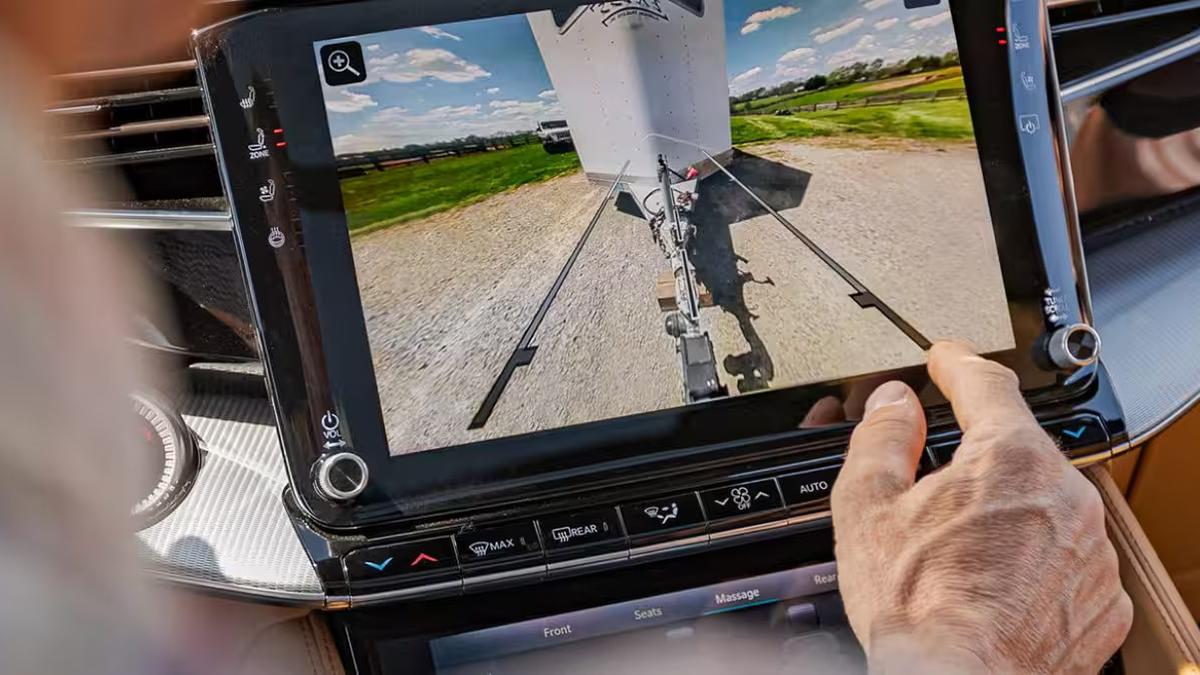 A close-up of a driver's hand interacting with a touchscreen displaying a rear view of a trailer, set in a scenic outdoor landscape.