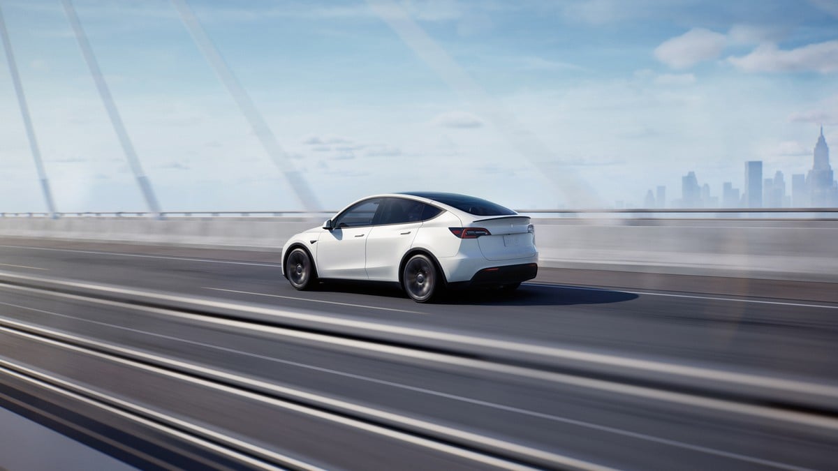 A sleek white Tesla Model Y speeding across a modern bridge, with a city skyline visible in the background.