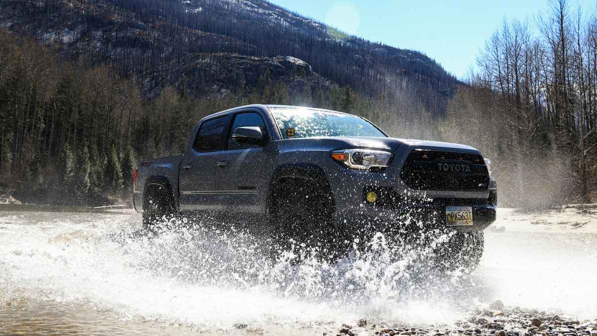 A 2024 Toyota Tacoma splashes through a shallow river, surrounded by lush trees and mountains under a bright blue sky.