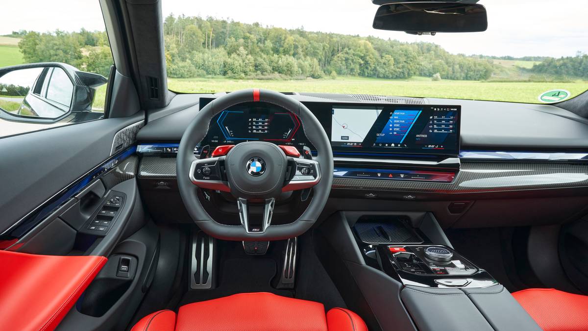 Interior view of a 2025 BMW M5 featuring a sleek dashboard, modern touchscreen displays, and sporty red seats, set against a green landscape.