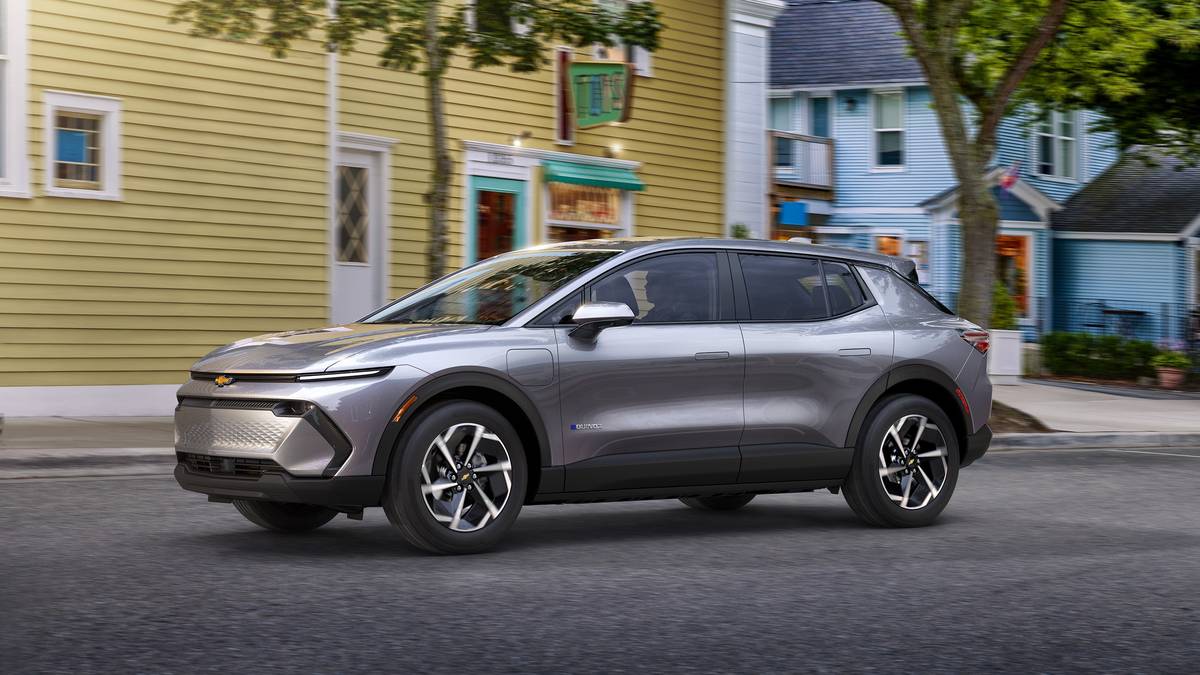 A sleek gray Chevrolet electric SUV drives past colorful storefronts in a charming, tree-lined street.