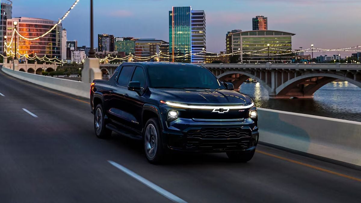 A black 2025 Chevrolet Silverado EV truck drives along a city road at dusk, illuminated by colorful city lights and a bridge in the background.
