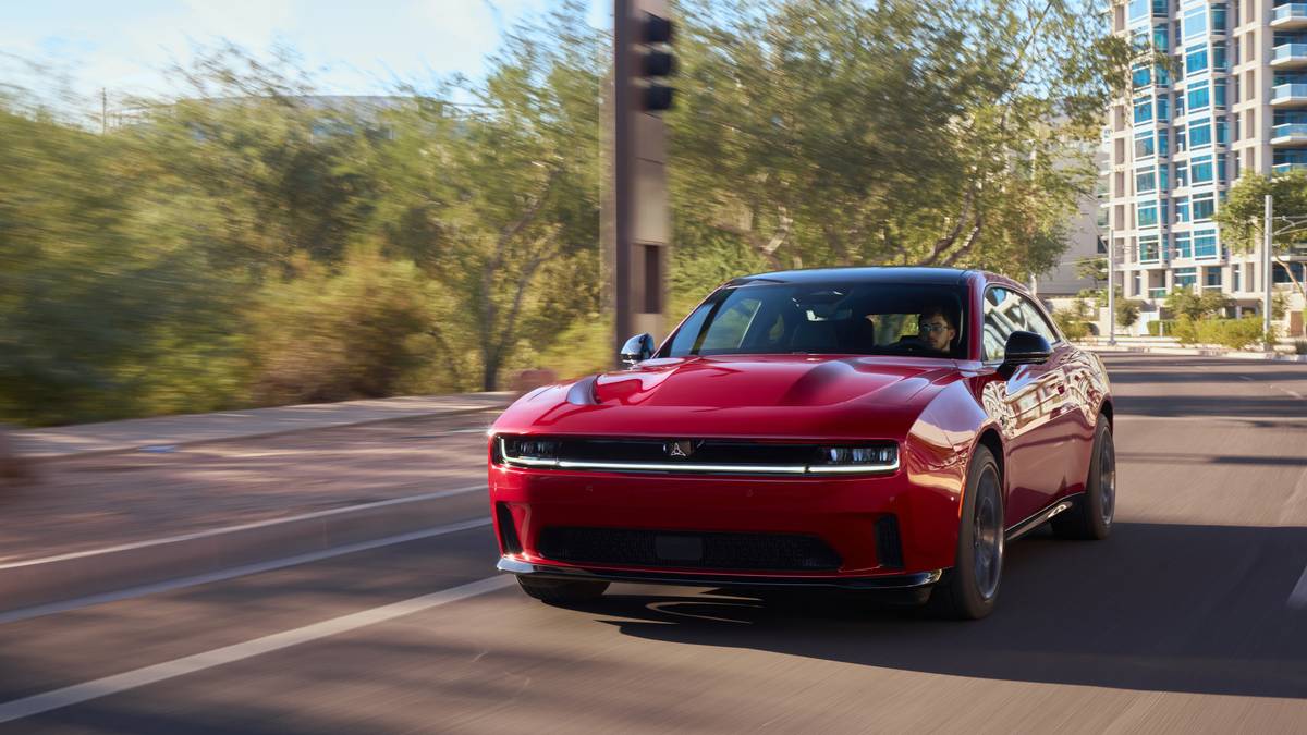 A sleek red sports car speeds down a tree-lined city street, framed by modern buildings under a clear blue sky.