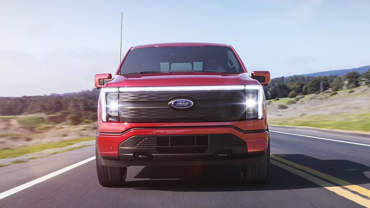 A red Ford truck drives through sandy terrain, kicking up dust, set against a vast desert landscape and rocky hills under a cloudy sky.