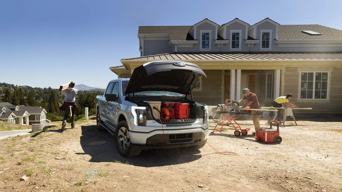 A silver 2025 Ford F-150 Lightning with an open hood sits on a construction site, while workers use tools and carry materials in front of a house.