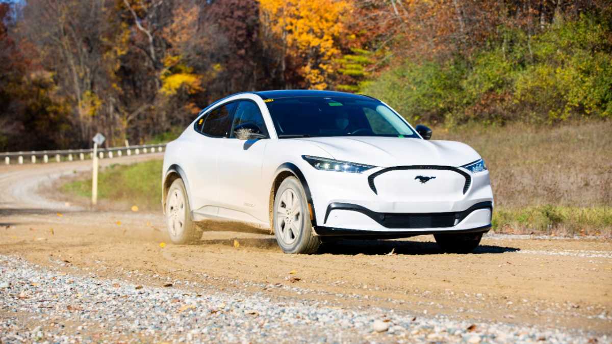 A white Ford Mustang Mach-E drives on a dusty gravel road surrounded by colorful autumn foliage.