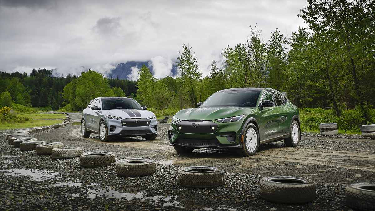 Two Ford Mustang Mach-E electric SUVs on gravel, one silver and one green, parked among tires with misty forest mountains in background