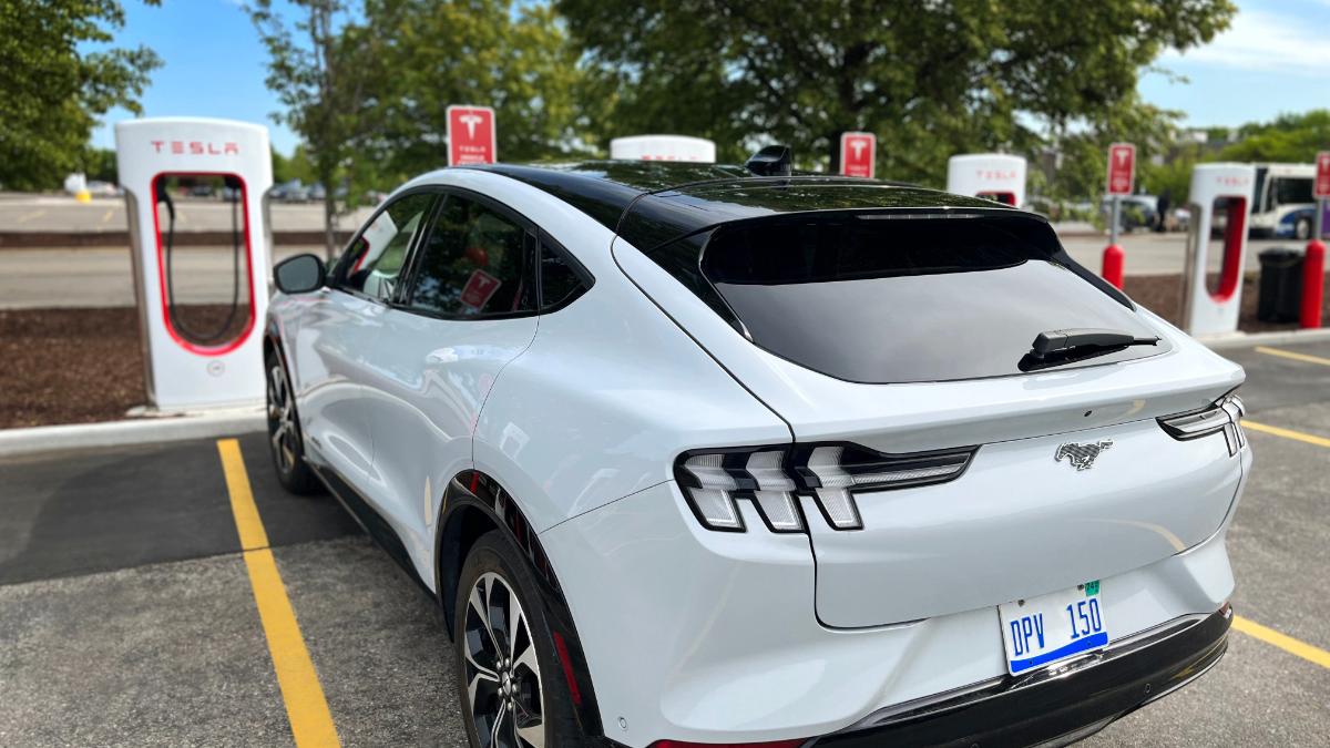 White Ford Mustang Mach-E electric SUV parked at Tesla charging station, sleek rear view with distinctive taillights and black roof.