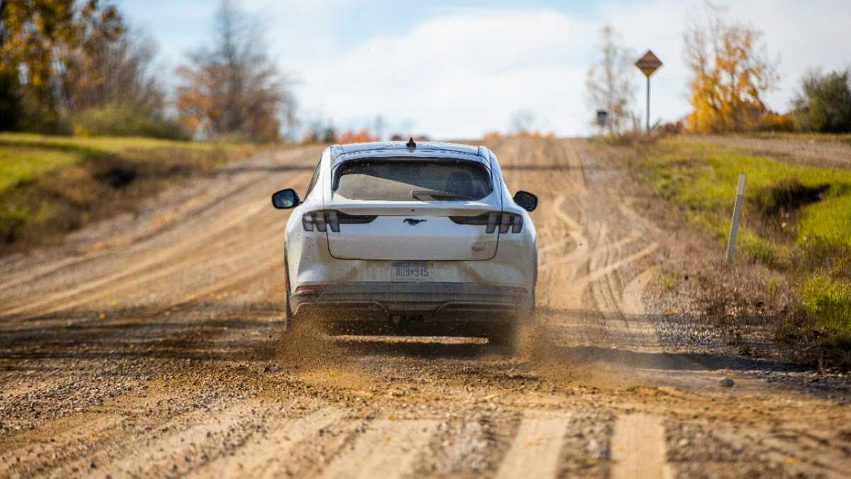A white SUV kicks up dust while driving on a gravel road lined with autumn foliage in the background.