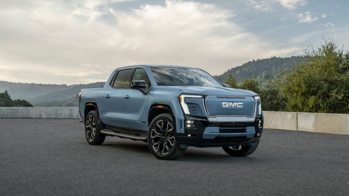 Blue GMC Sierra EV pickup truck parked on asphalt with forested mountain backdrop and cloudy sky