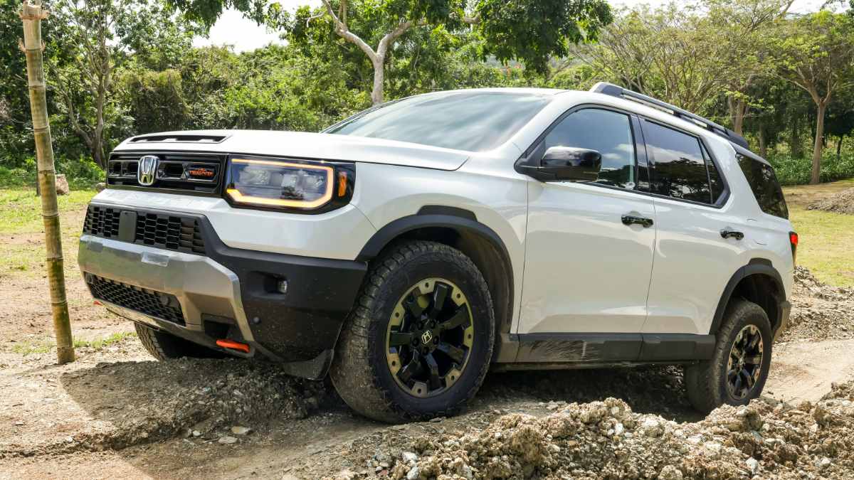 White 2025 Honda Passport SUV shown at three-quarter front angle on rugged terrain, featuring all-terrain tires, raised suspension, and distinctive LED headlights.