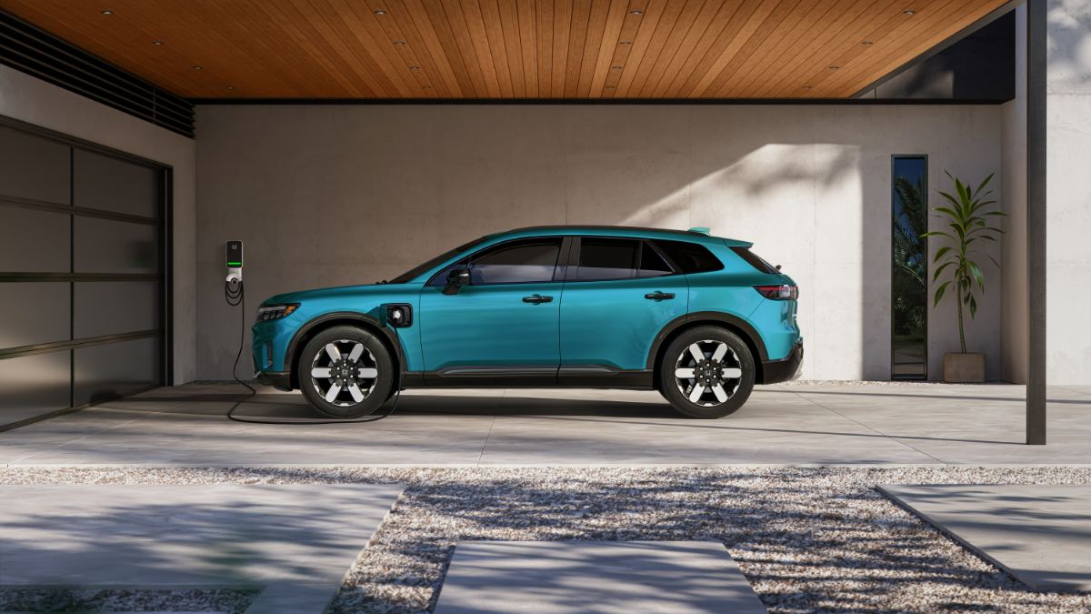 2024 Honda Prologue Elite electric SUV in turquoise, side view, charging at modern home garage with wood-paneled overhang, featuring distinctive two-tone wheels.