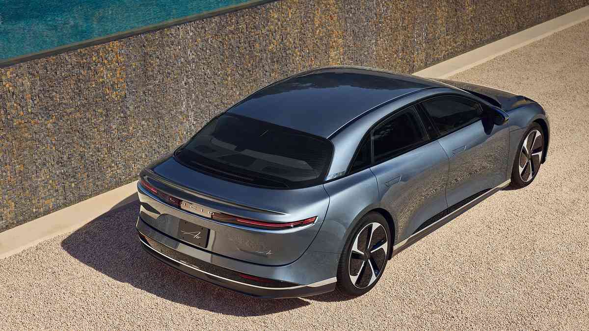 A silver 2025 Lucid Air electric sedan is parked beside a textured wall, overlooking a calm blue pool and surrounded by gravel.