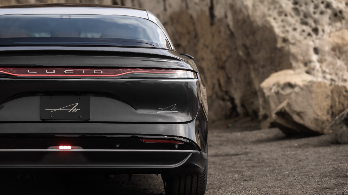 Black Lucid Air electric sedan rear view, illuminated red taillight, parked against rocky terrain backdrop