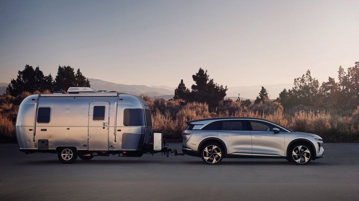 A Gray 2025 Lucid Gravity towing a sleek aluminum Airstream trailer against a backdrop of mountains and tall shrubs at sunset.