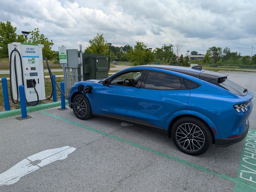 A blue electric vehicle charges at an EV fast charging station in a parking area, surrounded by greenery under a cloudy sky.