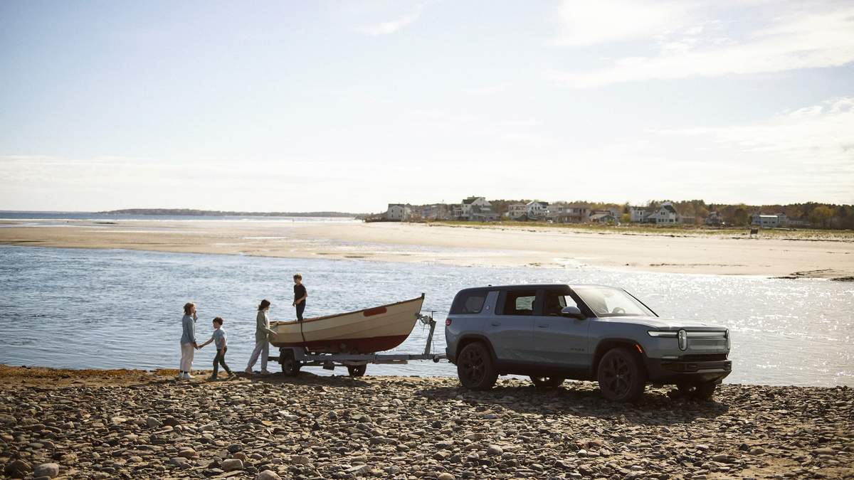 A family gathers near a river, unloading a boat from a vehicle on rocky shore, with homes lining the distant coastline under clear skies.