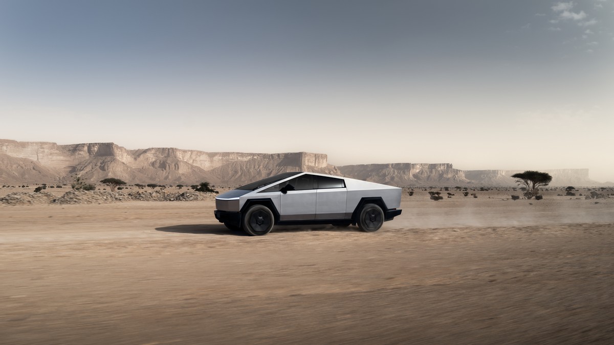 A silver Tesla Cybertruck drives on a dusty road in a desert landscape with rocky cliffs in the background.