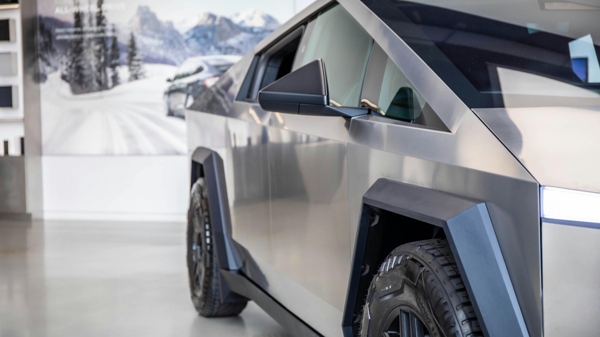A 2025 Tesla Cybertruck displays its metallic exterior and unique tire design in a modern showroom with snowy mountains in the background.