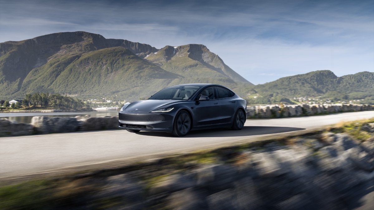 Black Tesla Model 3 driving on coastal road with dramatic mountain landscape in background