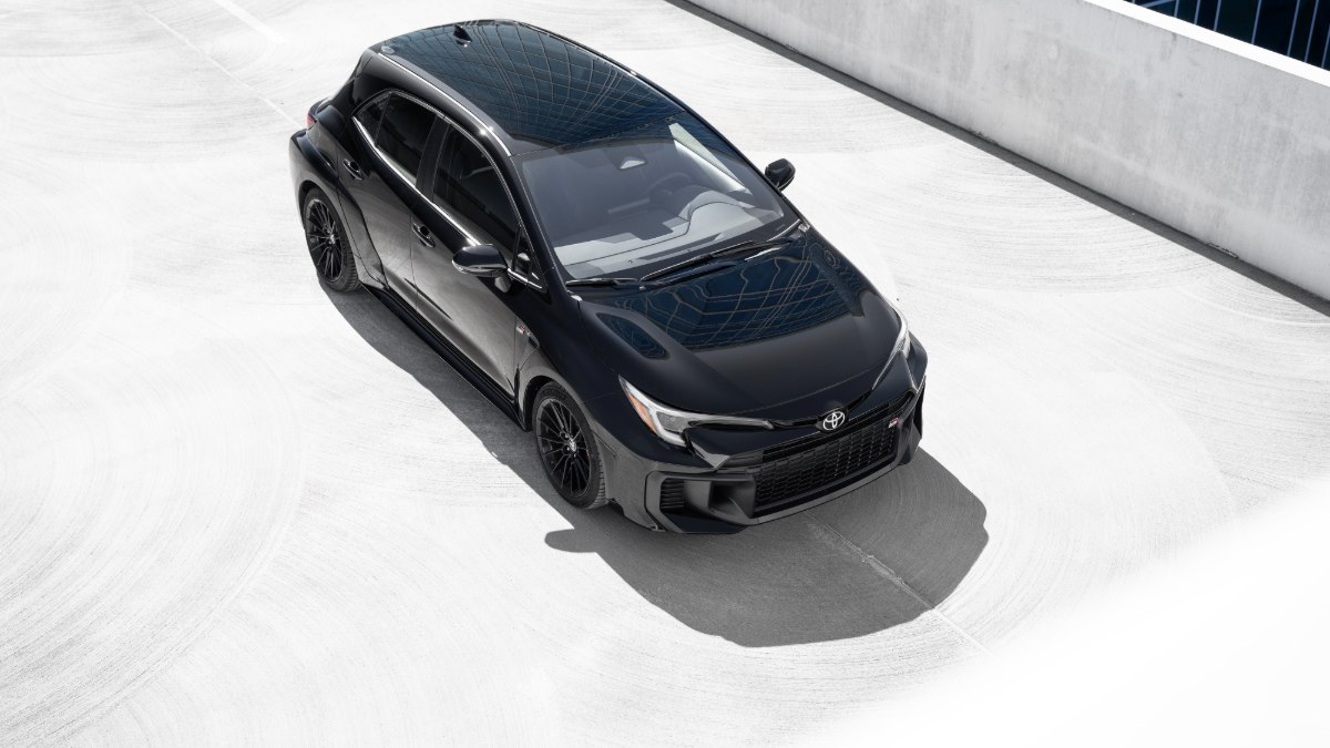 2023 Toyota GR Corolla in black, photographed from high overhead angle, featuring black alloy wheels, panoramic roof, and aggressive body styling, parked on light concrete surface.