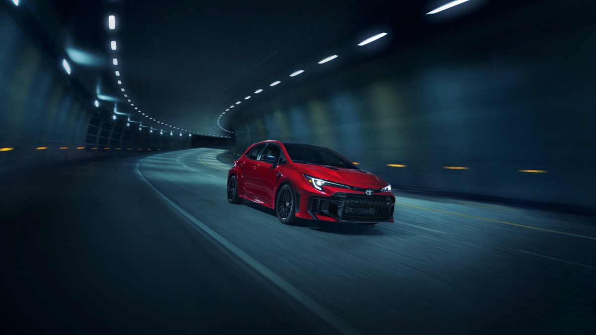 2024 Toyota GR Corolla in vibrant red, photographed at three-quarter front angle inside dimly lit tunnel with dramatic overhead lighting and curved road.