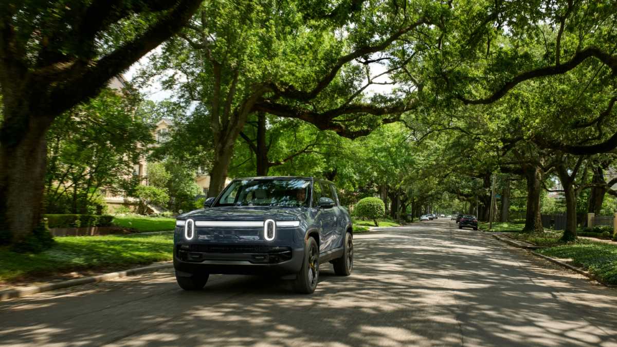 Electric truck driving on a tree-lined suburban street with dappled sunlight.