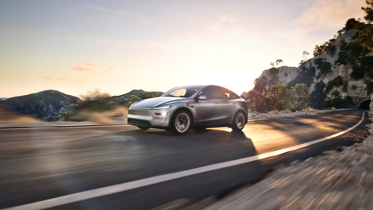 A sleek silver electric car drives along a winding road, with sunlit mountains in the background and a warm sunset glow.