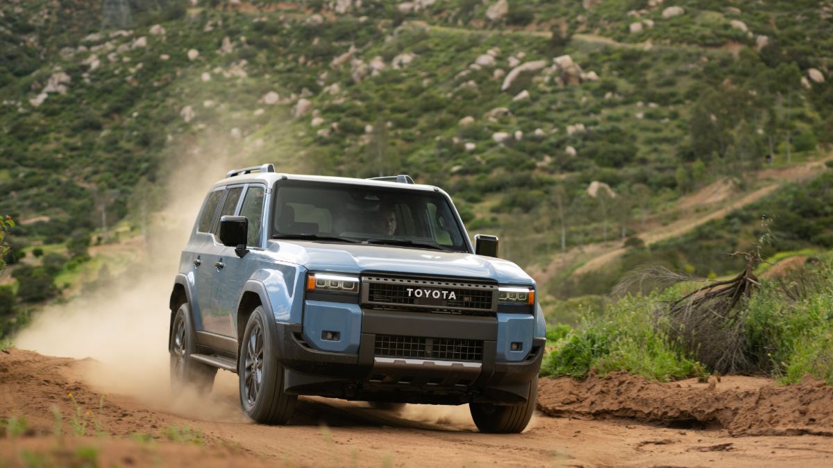 Blue Toyota SUV driving on a dusty dirt road surrounded by green hills and rocks.