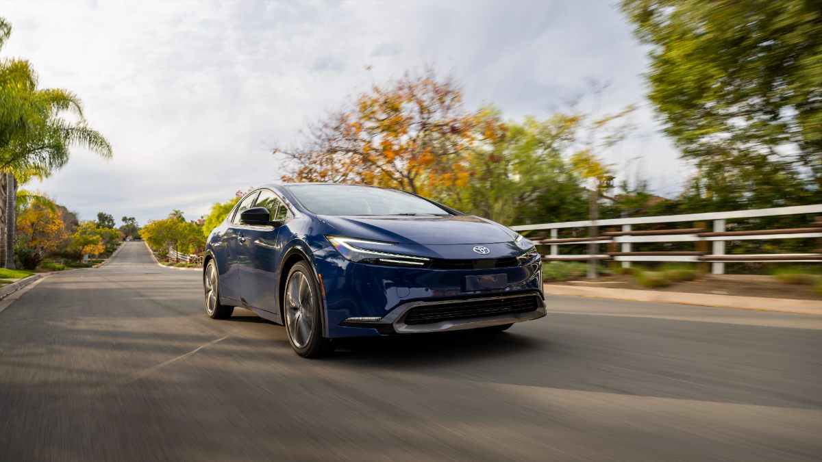 A blue Toyota Prius drives down a scenic tree-lined street, showcasing its sleek design against a backdrop of autumn foliage.
