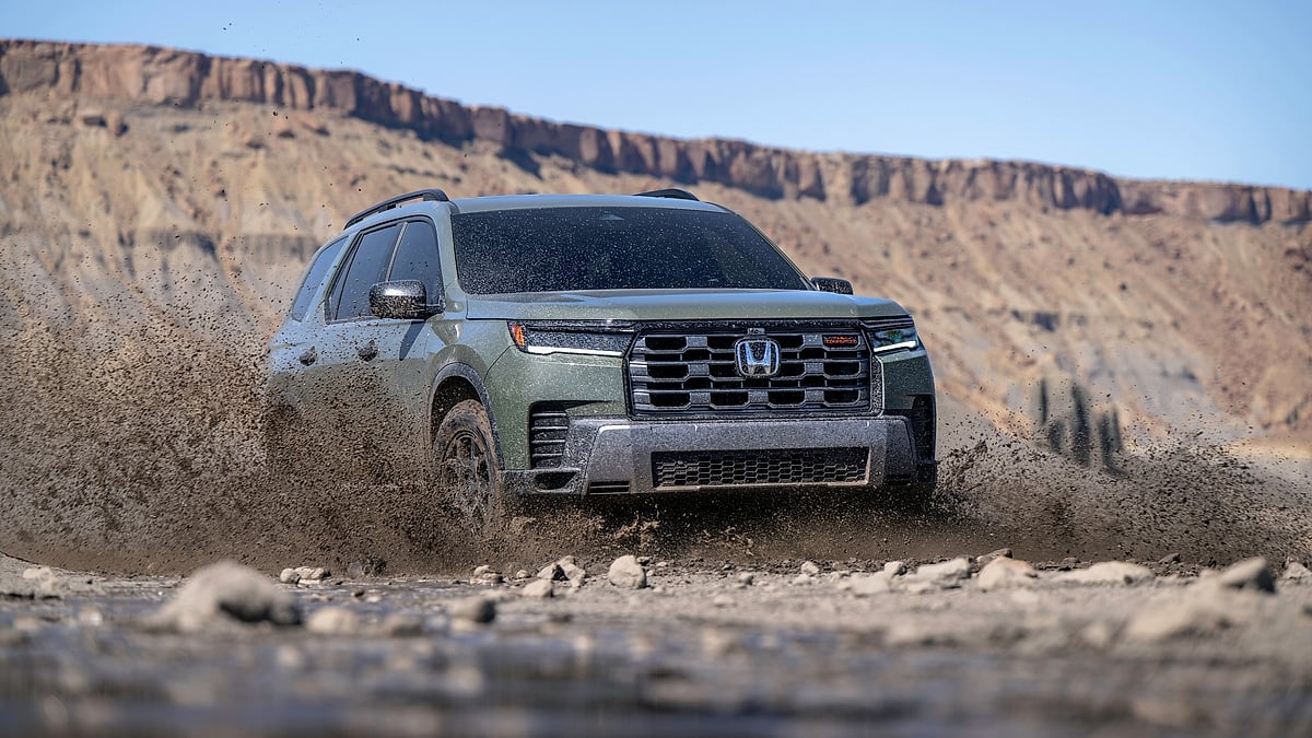2026 Honda Pilot TrailSport driving through muddy water on a desert off-road trail with cliffs in the background.