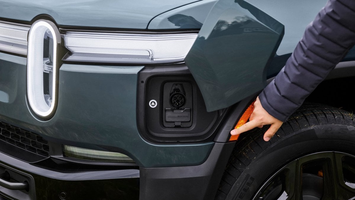 A close-up of an electric vehicle's charging port, with a person pointing at it, highlighting the integration with the vehicle's design.