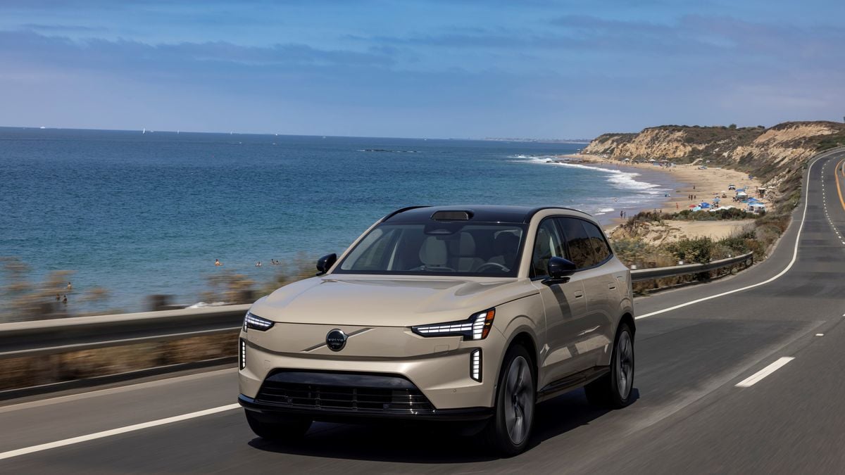 A beige 2025 Volvo EX90 drives along a coastal road with the ocean on one side and sandy beaches in the background under a clear blue sky.