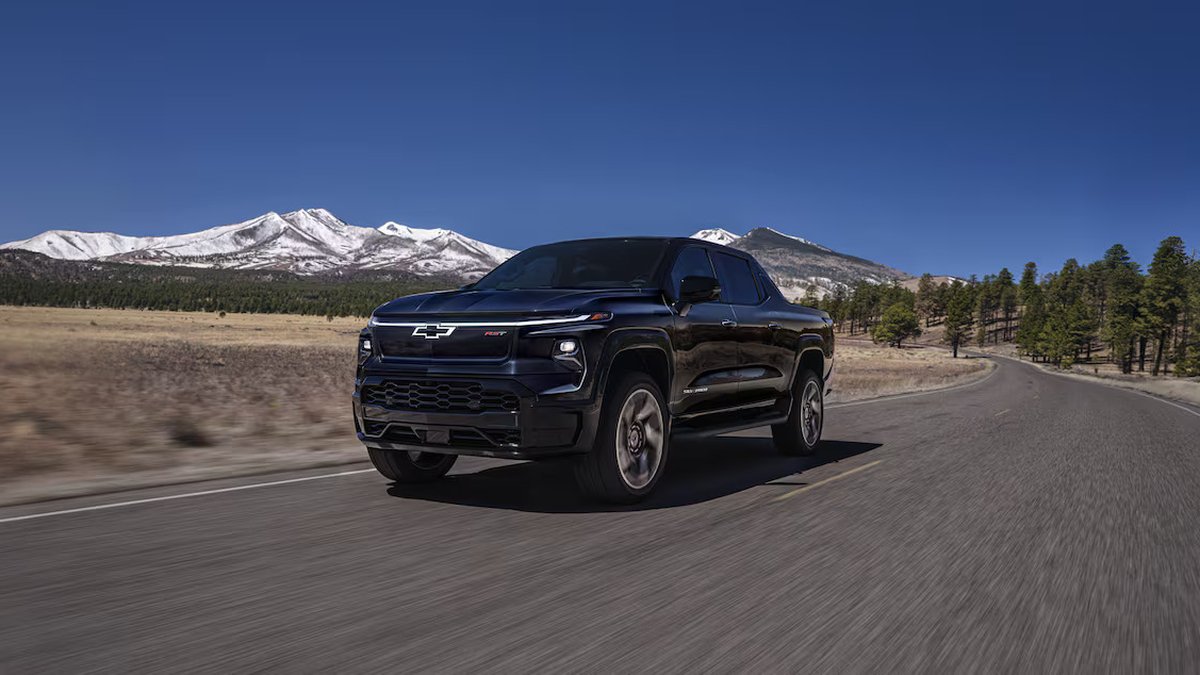 A black Chevrolet Silverado EV truck driving on a winding road, surrounded by snow-capped mountains and greenery under a clear blue sky.