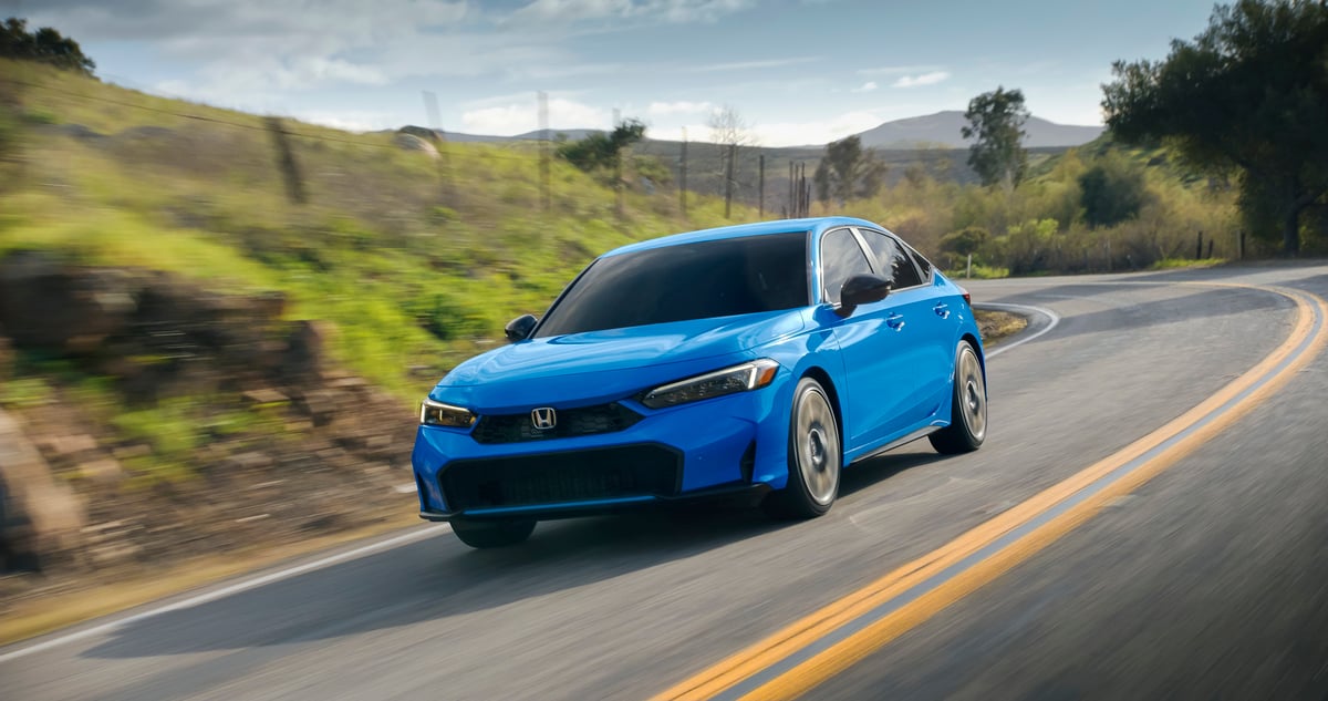 A vibrant blue Honda Civic Hybrid Sport Touring drives along a winding road, surrounded by lush green hills and a partly cloudy sky.