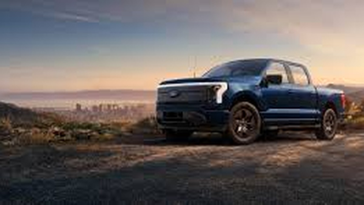 A blue Ford electric truck parked on a scenic overlook with a city skyline in the background during sunset.