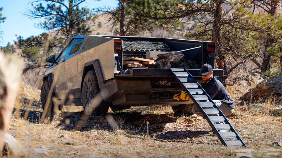 A rugged, 2025 Tesla Cybertruck loaded with wood and equipment sits in a wooded area, while a person uses a ramp to access the cargo.