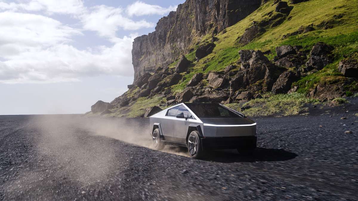 A 2025 Tesla Cybertruck kicking up dust on a rugged, black gravel road, surrounded by steep cliffs and lush greenery.