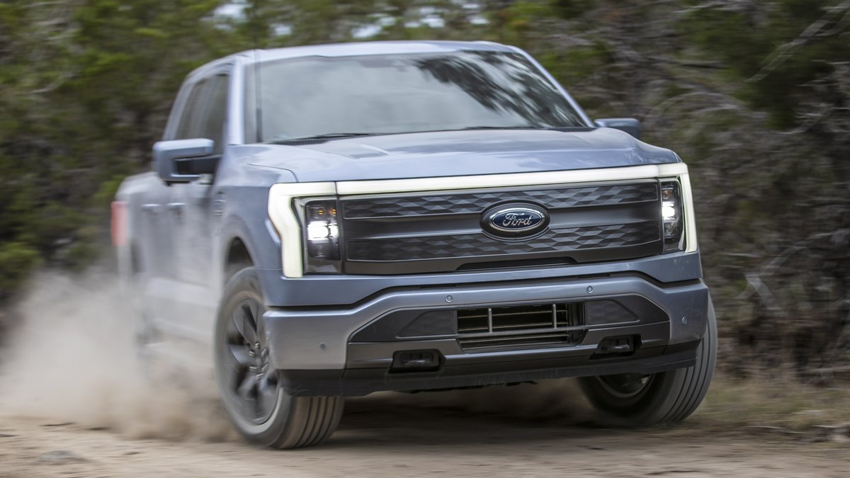 A Ford electric truck drives off-road, kicking up dust with a sleek design and bright headlights, set against a backdrop of green foliage.