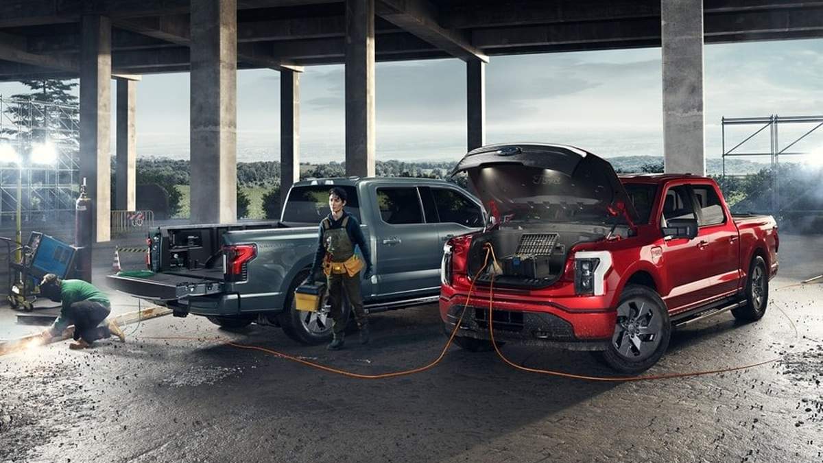 Two trucks, one gray and one red, are parked under a structure; a worker is repairing the red truck's engine while another works nearby.