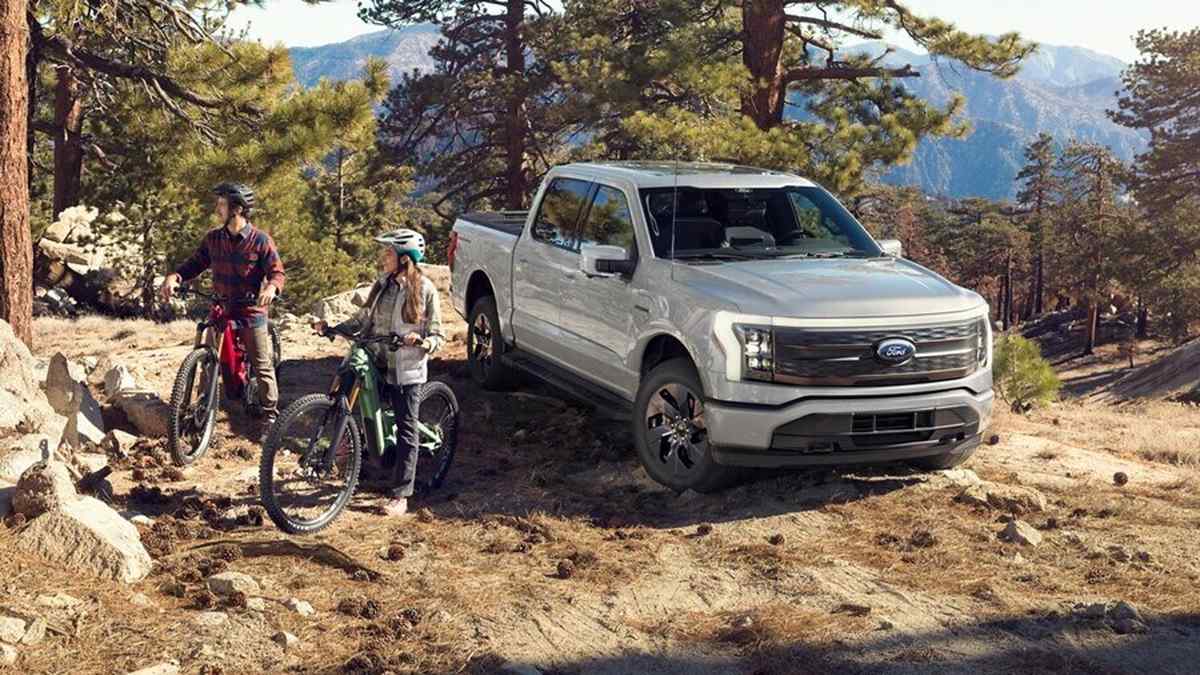 A silver Ford truck parked on a rugged mountain trail, with two cyclists beside it surrounded by tall pine trees and rocky terrain.