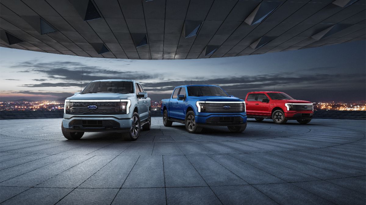 Three Ford F-150 Lightning electric trucks in white, blue, and red under dramatic architectural ceiling at dusk