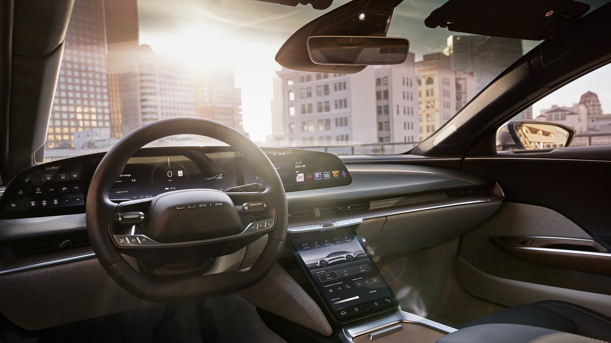 Interior view of a2025 Lucid Air Touring, featuring a modern dashboard, a sleek steering wheel, and a cityscape backdrop at sunrise.